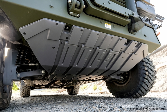 The image shows the underside of a military vehicle, highlighting its protective underbody plating and suspension components.